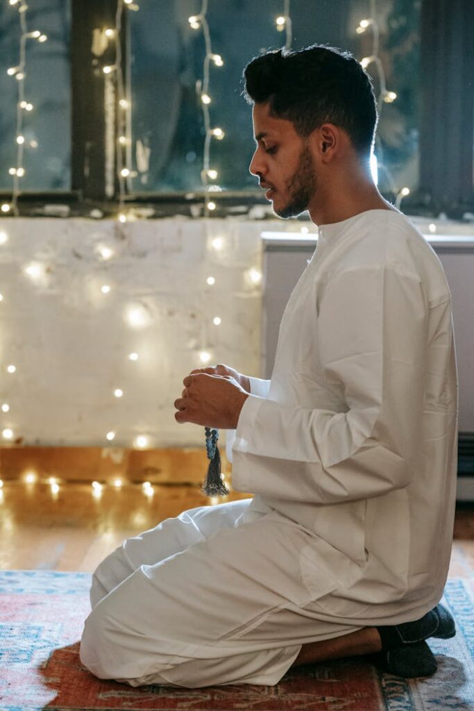 Side view of a Muslim man in traditional clothing praying indoors with string lights in the background.