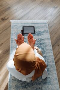 A woman in a hijab prays on a rug with a tablet nearby, in a peaceful indoor setting.