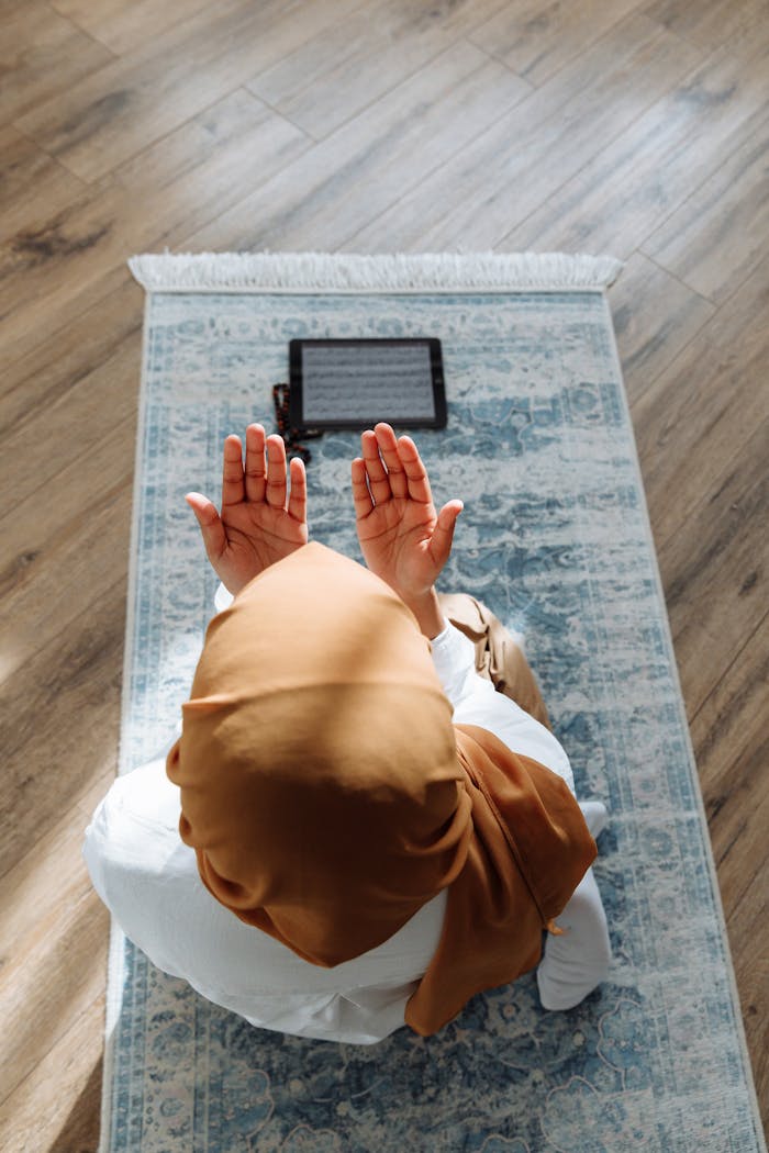A woman in a hijab prays on a rug with a tablet nearby, in a peaceful indoor setting.
